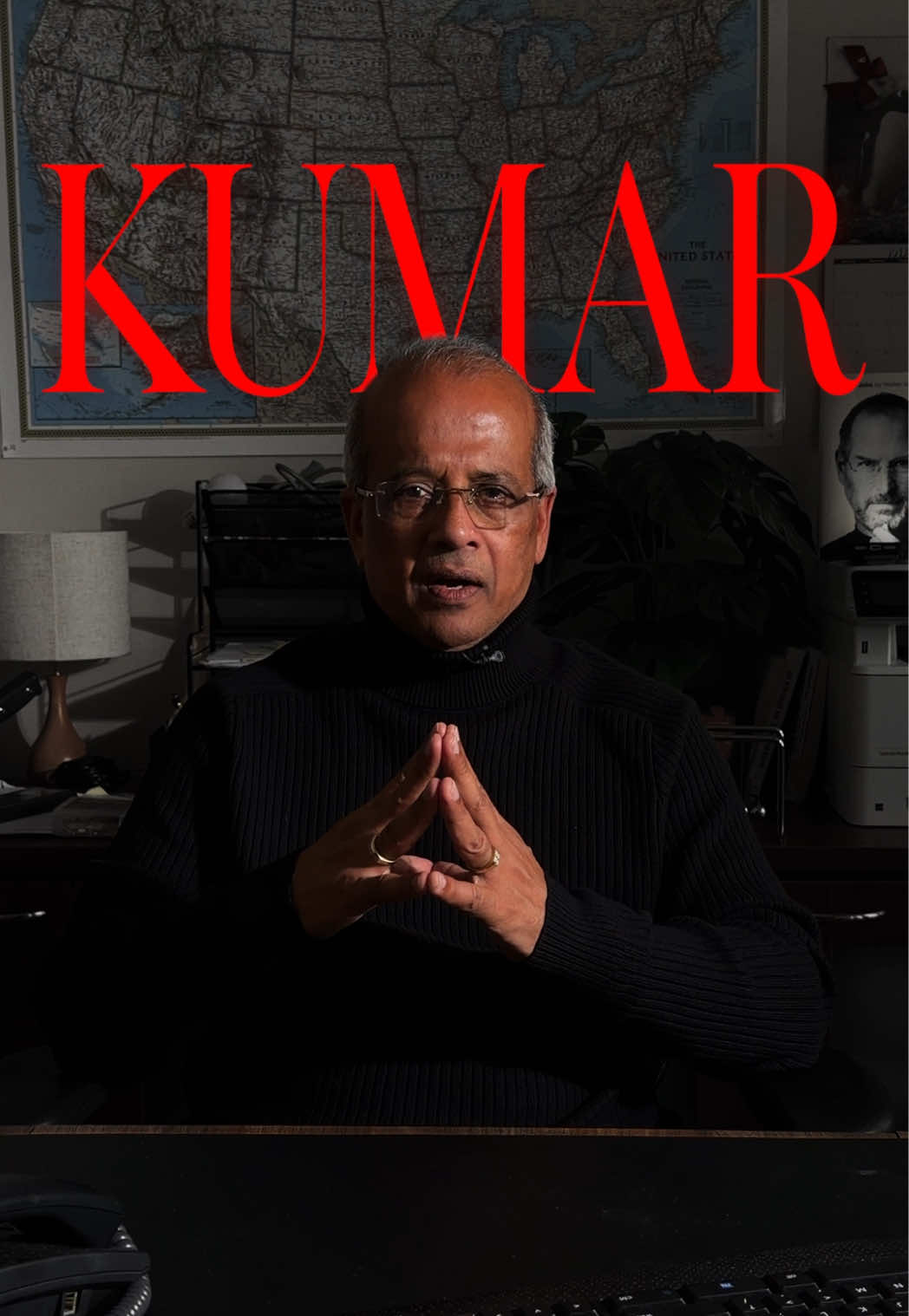 Kumar steepling his fingers behind his desk under a giant red KUMAR title card